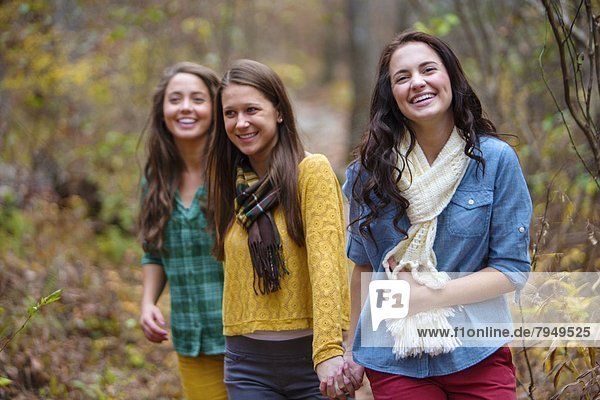 Three young women smile & hold hands while walking down a trail.