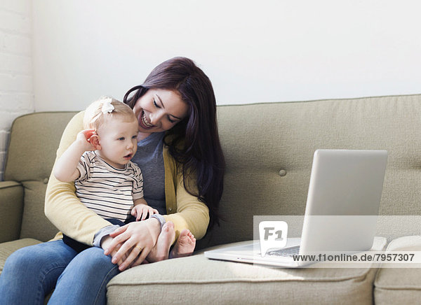 Woman and baby girl (12-17 months) sitting on sofa and using laptop
