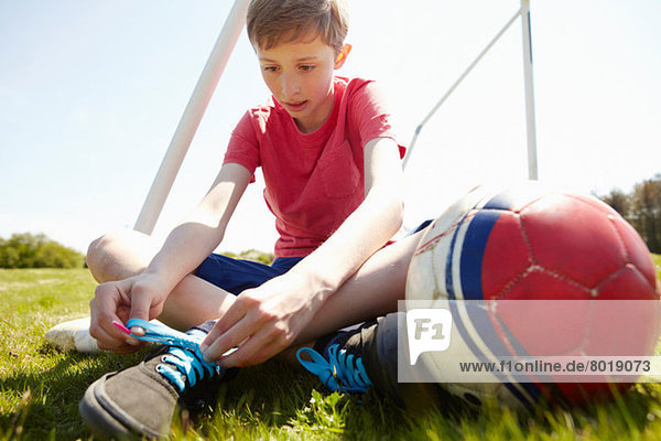 Junge sitzt auf dem Feld und bindet Schnürsenkel.