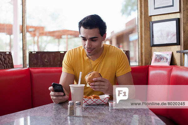 Young man texting on mobile phone and eating fast food in diner