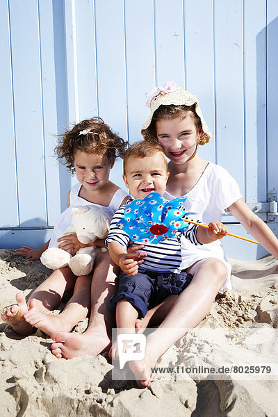 Two girls and a toddler are sitting on the beach in front of a beach hut.