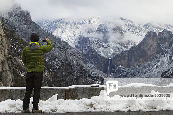 Man wearing a green down jacket as seen from the back taking a picture of yosemite valley and bridal veil falls Yosemite california united states of america