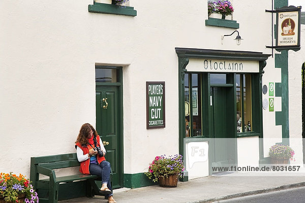 A woman sits on a bench outside a shop looking at her camera Ballyvaughan county clare ireland