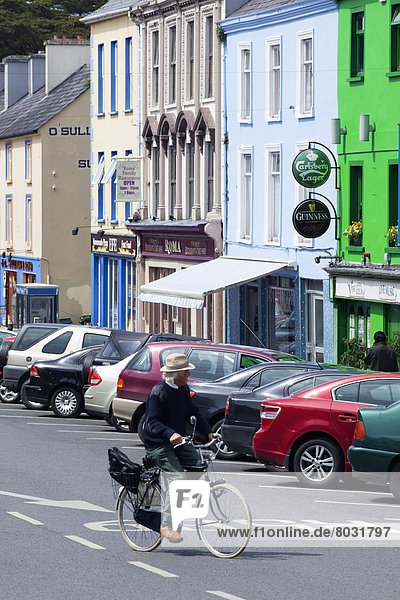 A cyclist rides down main street going by parked cars and colourful buildings Kenmare county kerry ireland