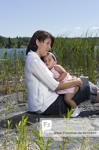 Native Aboriginal Mother Sitting With Her One Year Old Daughter On A Rock Beside A Lake With Reeds In Shoal Lake  Ontario  Canada.