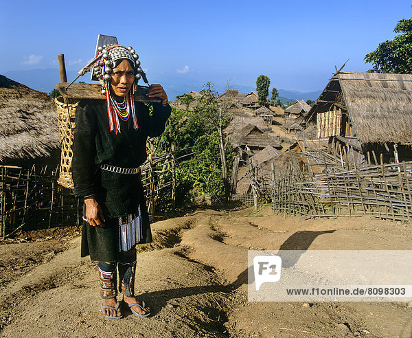 Akha-Frau in traditioneller Kleidung  mit Rückentrage  in einem Bergdorf mit Bambushütten