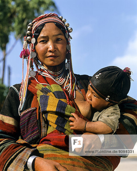 Junge Akha-Frau stillt Baby  mit traditioneller Kleidung und Kopfbedeckung