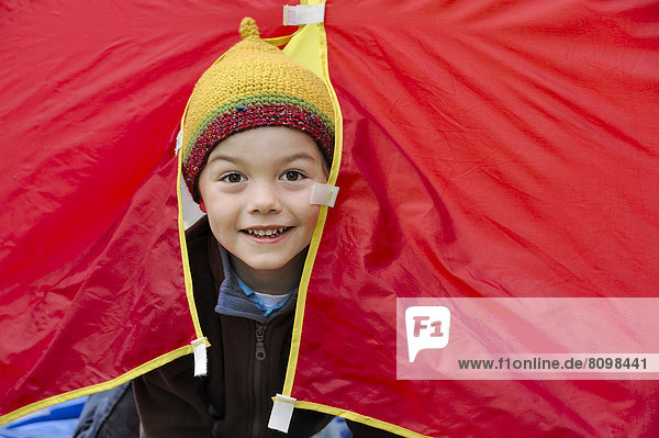 Ein Junge mit einer Mütze schaut fröhlich aus einem roten Zelt heraus