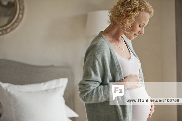 Lächelnde schwangere Frau berührt den Bauch im Schlafzimmer