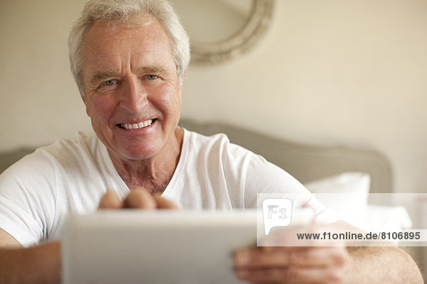 Porträt eines lächelnden älteren Mannes mit digitalem Tablett im Schlafzimmer