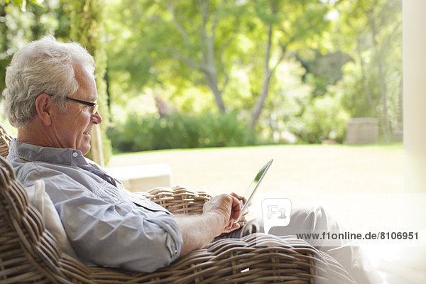 Senior Mann mit digitalem Tablett im Korbsessel auf der Veranda