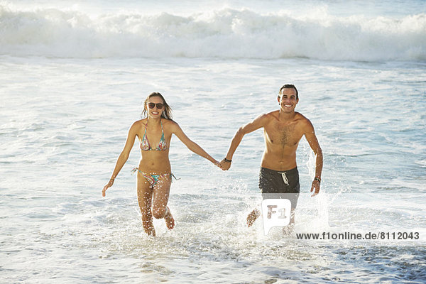 Porträt eines glücklichen Paares  das Händchen hält und im Ozean rennt