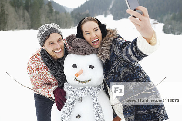 Enthusiastic couple taking self-portrait with snowman in snowy field