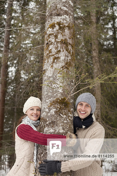 Portrait of happy couple hugging tree trunk in woods