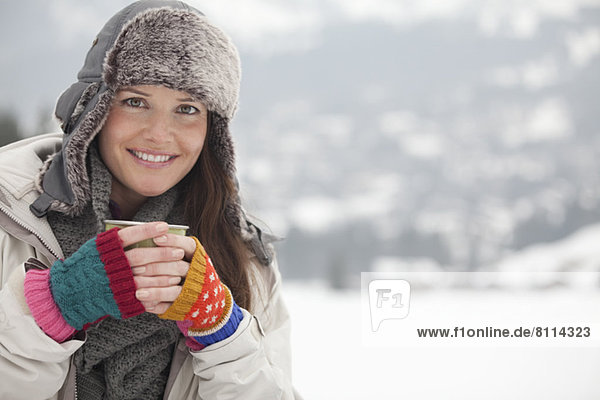 Portrait of happy woman in fur hat drinking coffee in snowy field