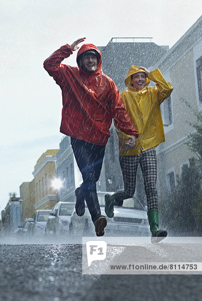 Glückliches Paar in Regenmänteln  die im Regen die Straße hinunterlaufen