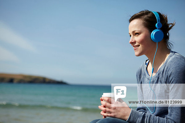 Junge Frau an der Küste mit Kaffee und Kopfhörer