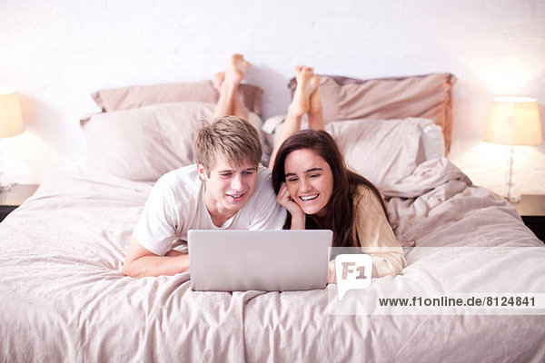 Junges Paar liegt auf dem Bett und schaut auf den Laptop.