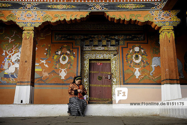 Eine junge Frau sitzt vor dem reich verzierten Eingang eines Nebentempels im Dzong von Paro  der Burgfestung Rinpung Dzong