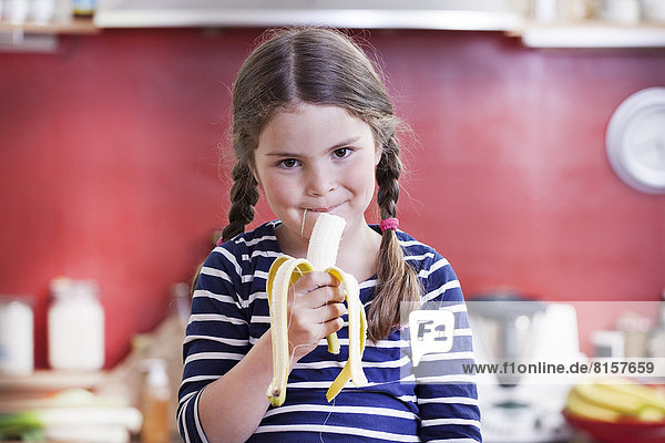 Germany  North Rhine Westphalia    Portrait of girl eating banana in kitchen