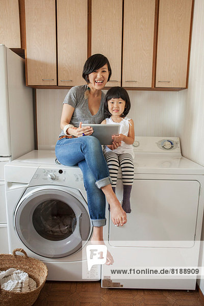 Mother and daughter sitting on washing machine using tablet