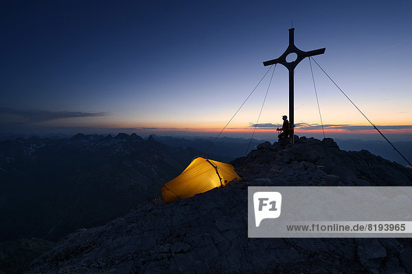 Mountaineer with a tent at the summit cross on Großer Krottenkopf Mountain at dusk