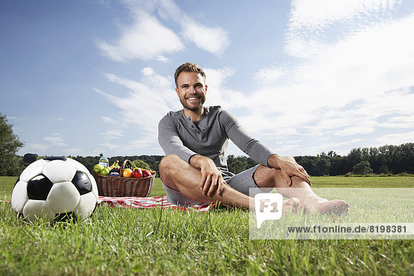 Mann auf Decke sitzend mit Picknickkorb und Fußball