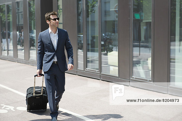 Businessman with sunglasses pulling luggage  walking in city