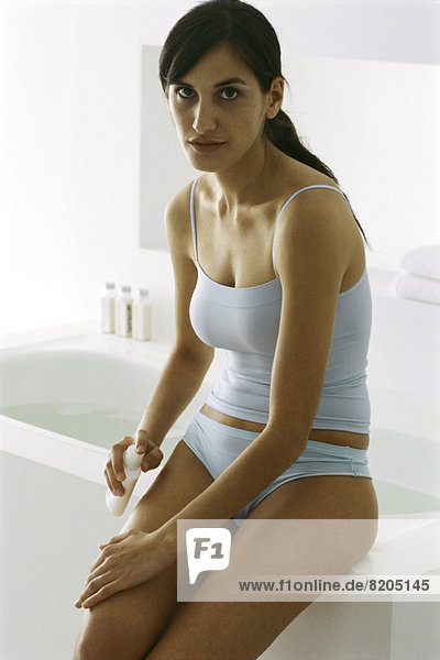 Frau sitzt auf Badewannenrand  setzt Feuchtigkeitscreme auf das Bein  schaut in die Kamera