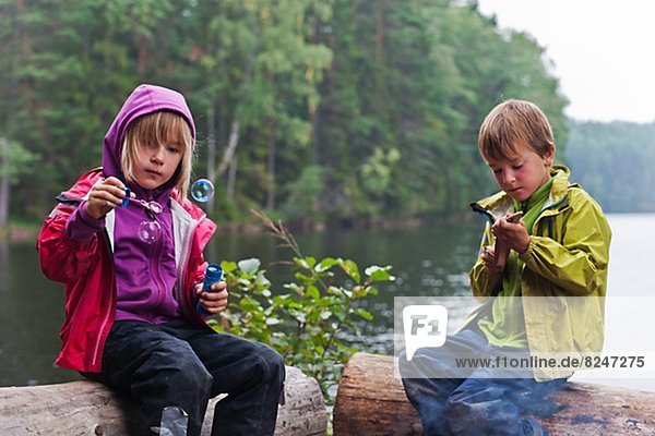 Boy and girl sitting on logs by lake  girl blowing bubbles
