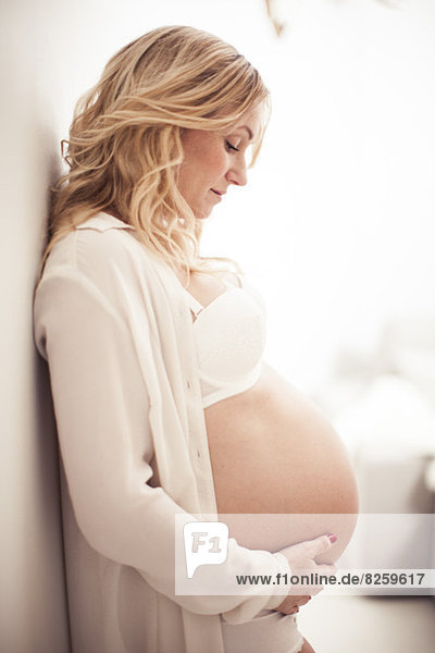 Side view of pregnant woman touching abdomen while leaning on wall at home