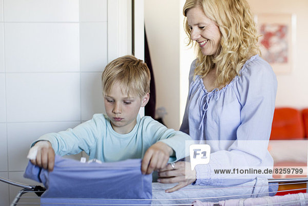 Lächelnde Mutter schaut auf den Sohn und trocknet die Wäsche auf dem Gestell.
