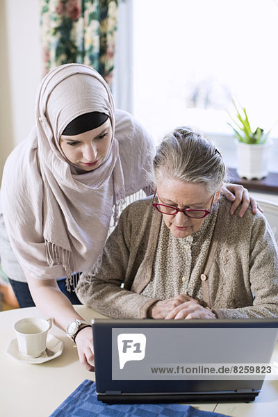 Seniorin mit häuslicher Betreuerin mit Laptop am Tisch