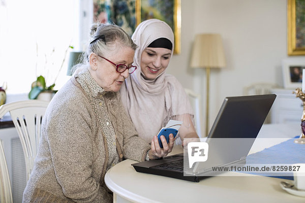 Junge Heimpflegerin mit älterer Frau  die mit dem Laptop zu Hause in die Bank geht.