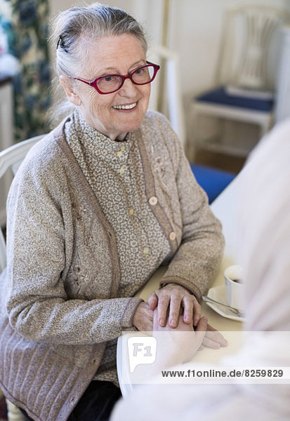 Lächelnde Seniorin hält die Hand der Hauspflegerin bei Tisch
