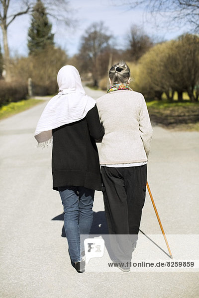 Rückansicht der älteren Frau und der Betreuerin im Haus  die Arm in Arm auf der Straße gehen