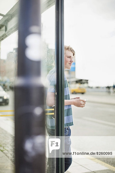 Junger Mann mit Handy im Stehen an der Bushaltestelle