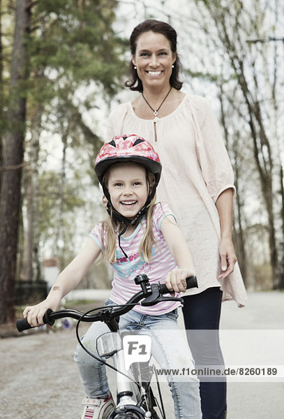 Portrait of happy girl riding bicycle while mother standing behind her on road
