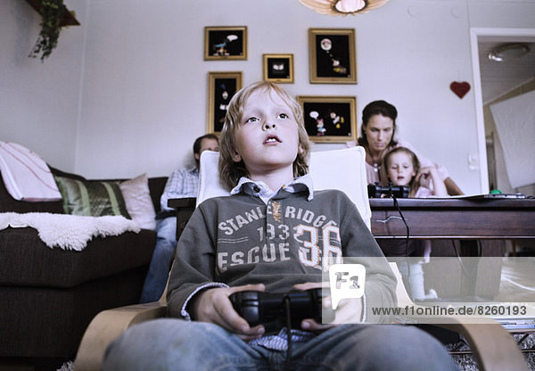 Kleiner Junge spielt Videospiel zu Hause mit Familie im Hintergrund