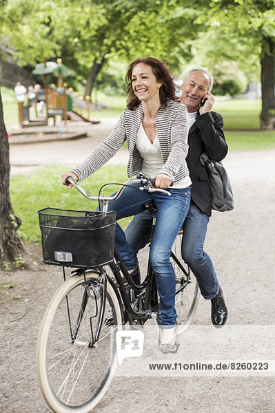 Glückliche Geschäftsfrau auf dem Fahrrad  während die Kollegin mit dem Handy auf dem Rücksitz sitzt.