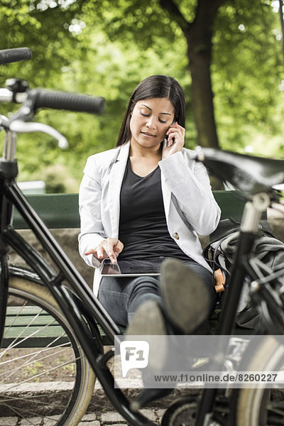 Geschäftsfrau mit digitalem Tablett und Handy auf Parkbank mit Füßen auf dem Fahrrad