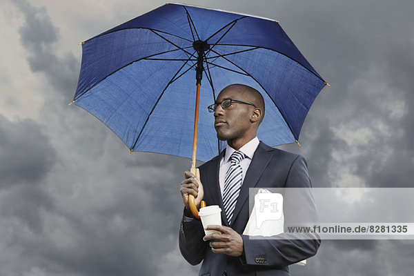 Businessman holding umbrella  disposable coffee cup and newspaper under a moody sky