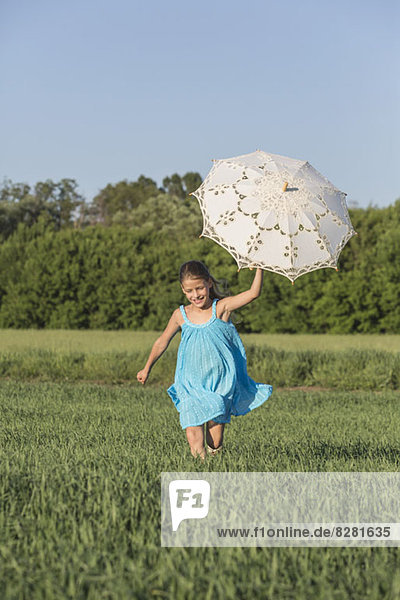 Ein junges Mädchen hält einen Regenschirm hoch  während es im Sommer durch ein Feld rennt.