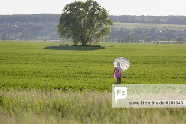 Ein junges Mädchen geht durch ein Feld und benutzt einen Regenschirm als Sonnenschutz.