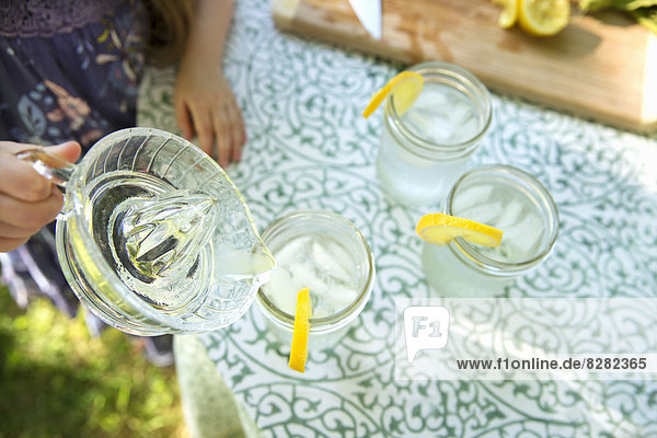 Limonade machen. Überkopfaufnahme von Limonadengläsern mit einer frischen Zitronenscheibe an der Glaskante. Ein Kind  das das Getränk aus einem Krug gießt.