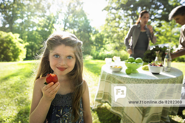 Ein junges Mädchen mit einer großen frischen  biologisch produzierten Erdbeere. Zwei Erwachsene neben einem runden Tisch.