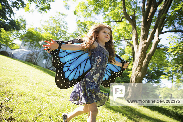 Ein Kind rennt vor einem Bauernhaus über den Rasen  mit großen reißenden blauen Schmetterlingsflügeln und mit ausgestreckten Armen.