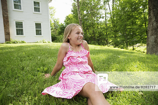 Ein junges Mädchen in einem rosa gemusterten Sonnenkleid  das auf dem Gras unter den Bäumen in einem Bauerngarten sitzt.
