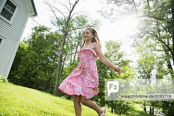 Ein junges Mädchen in einem rosa gemusterten Sonnenkleid  das in einem Bauerngarten unter den Bäumen über das Gras läuft.