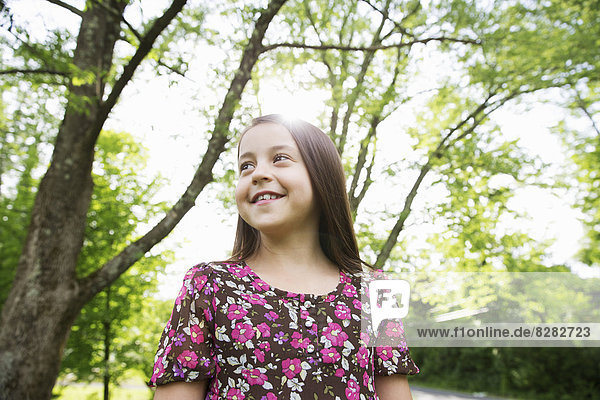Ein junges Mädchen in einem gemusterten Sommerkleid  unter dem Schatten von Bäumen in einem Bauerngarten.